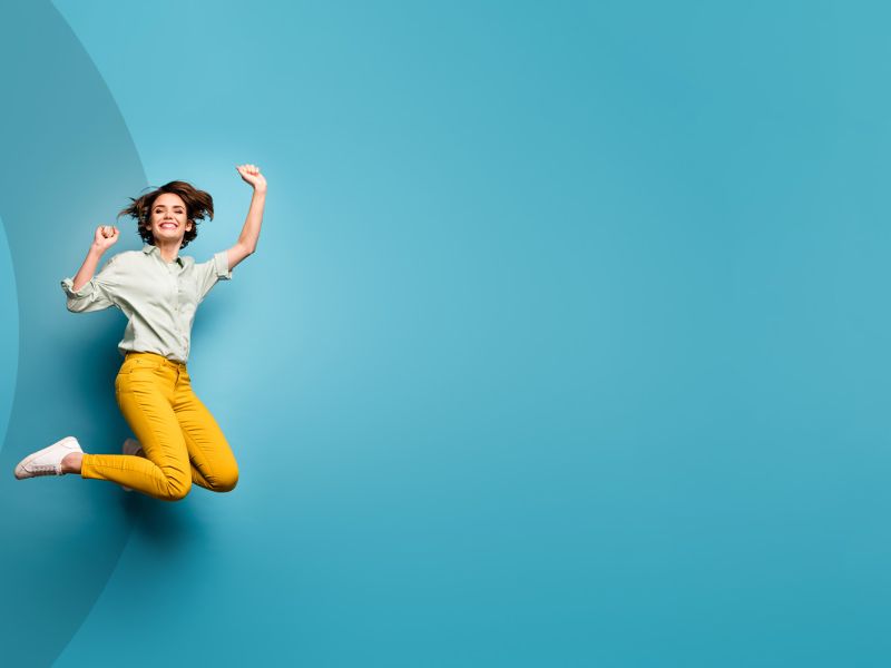 Eine Person springt freudig in einer hellblauen Umgebung, trägt ein grünes Hemd und eine leuchtend gelbe Hose und strahlt Begeisterung und Energie aus.