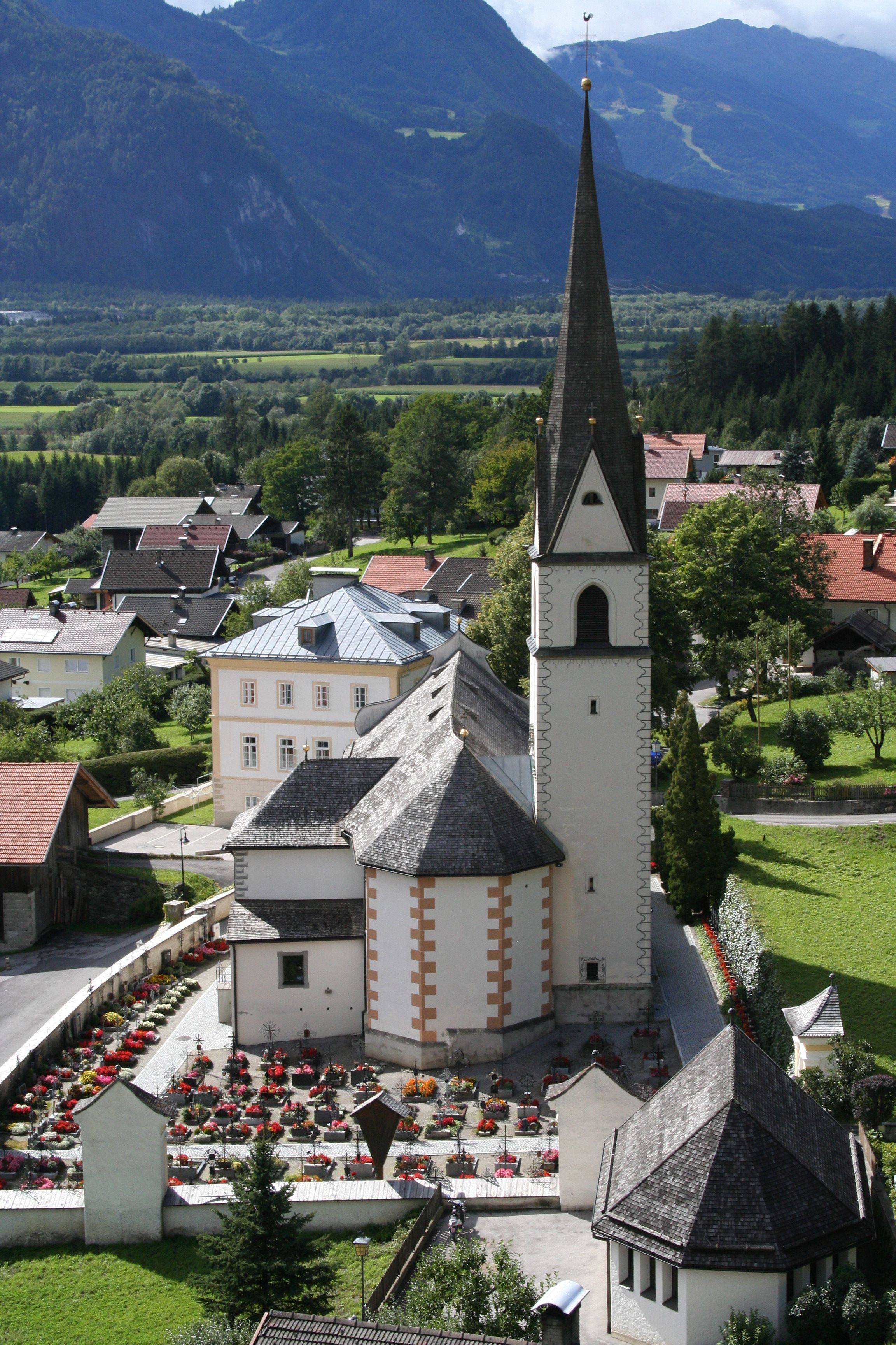 Eine malerische Kirche mit einem hohen Kirchturm steht vor dem Hintergrund eines blauen Himmels und grüner Berge, umgeben von üppigem Gras und Bäumen.