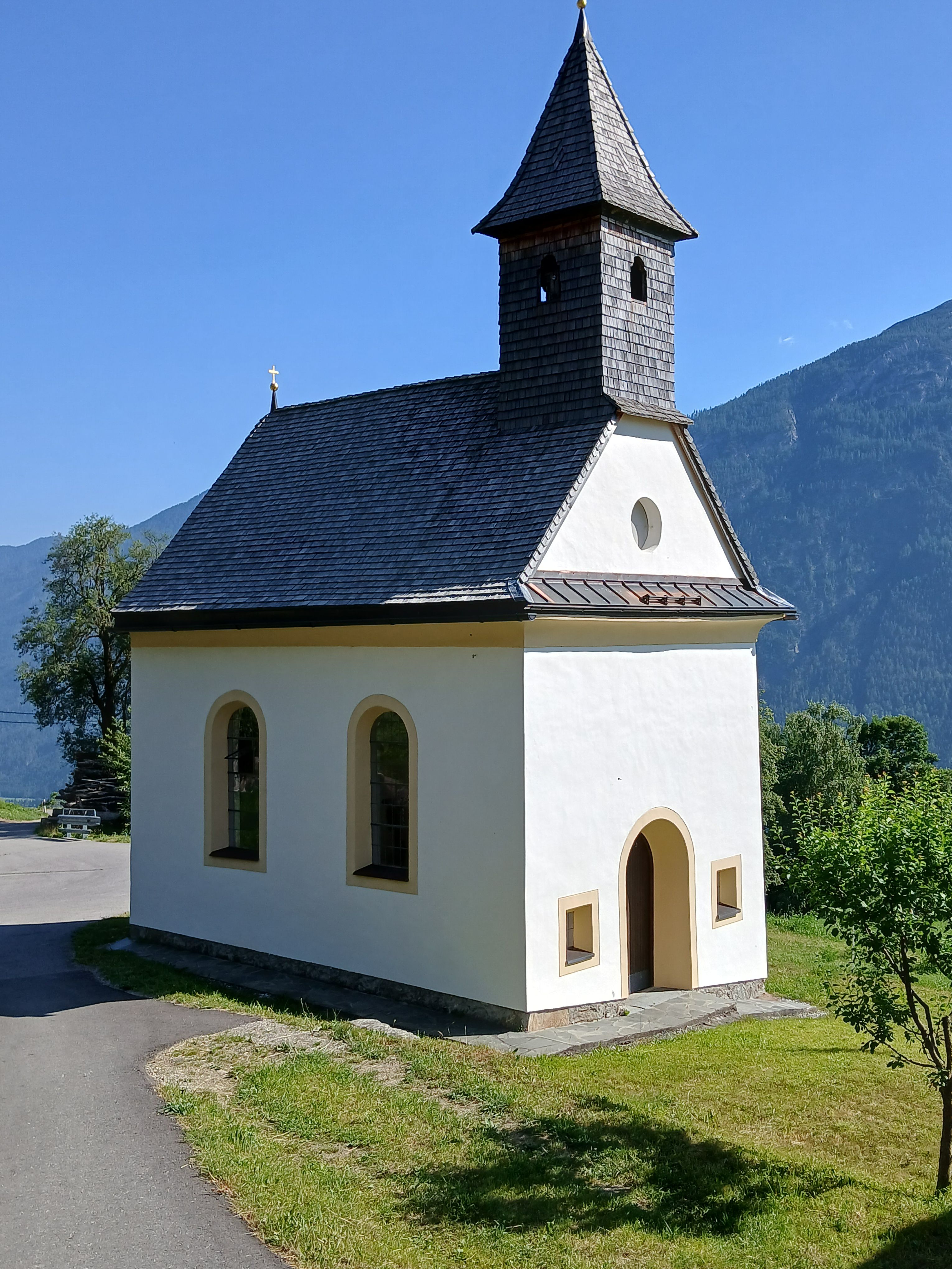 Kleine helle Kapelle mit spitzem Turm steht auf einer Wiese vor einer Bergkulisse unter blauem Himmel.
