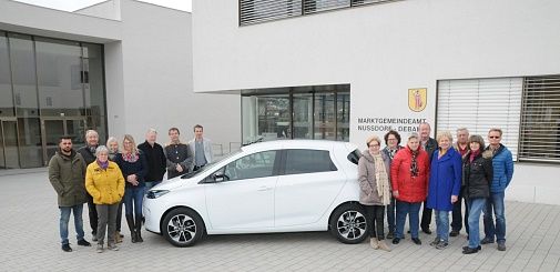 Eine Gruppe unterschiedlicher Personen steht neben einem weißen Auto vor dem Gebäude der Marktgemeinde Nussdorf-Debant.