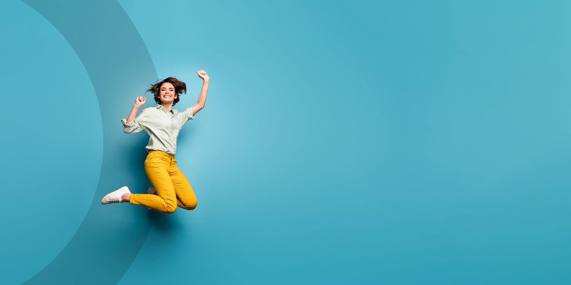 Eine Person springt freudig in einer hellblauen Umgebung, trägt ein grünes Hemd und eine leuchtend gelbe Hose und strahlt Begeisterung und Energie aus.