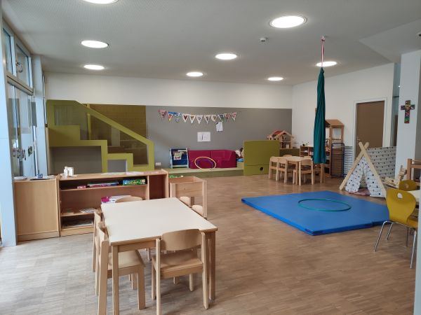 Spielbereich des Kindergartens, im Vordergrund steht ein Kindertisch, im Hintergrund ist eine Turnmatte und eine Spielecke